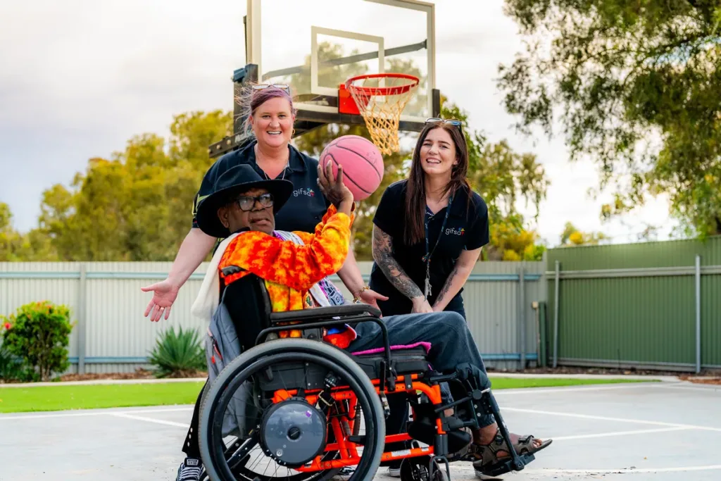 A participant in a wheelchair shooting a basketball, cheered on by two GIFSA support workers