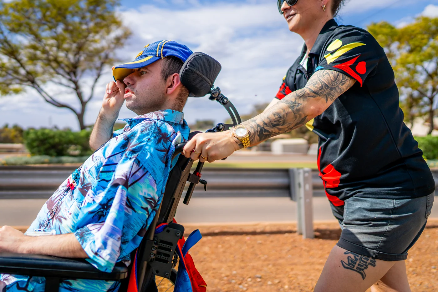 A GIFSA support worker pushing a participant's wheelchair along a path beside the road