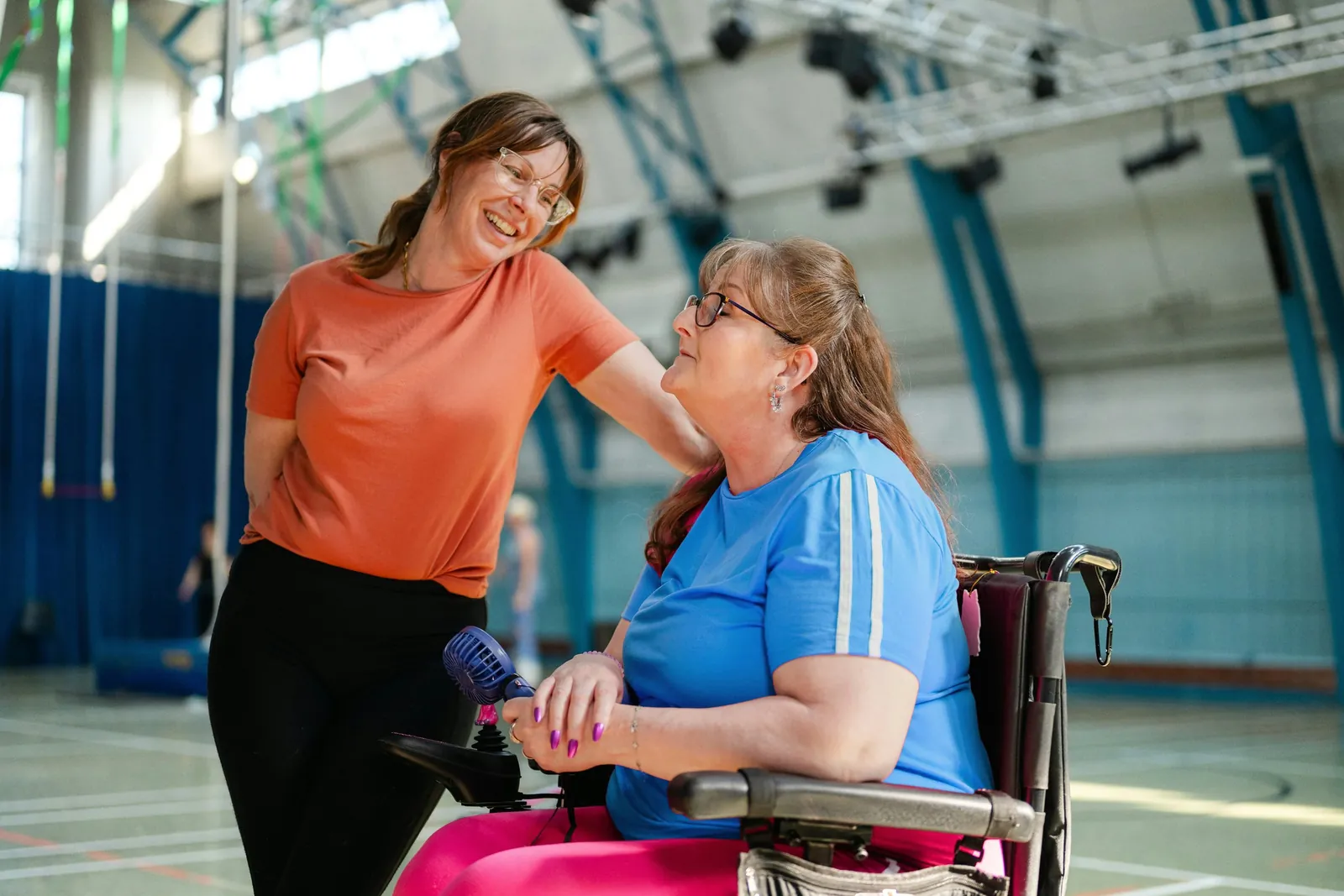 Two women laughing together in a community sports hall, one resting a hand on the other's shoulder