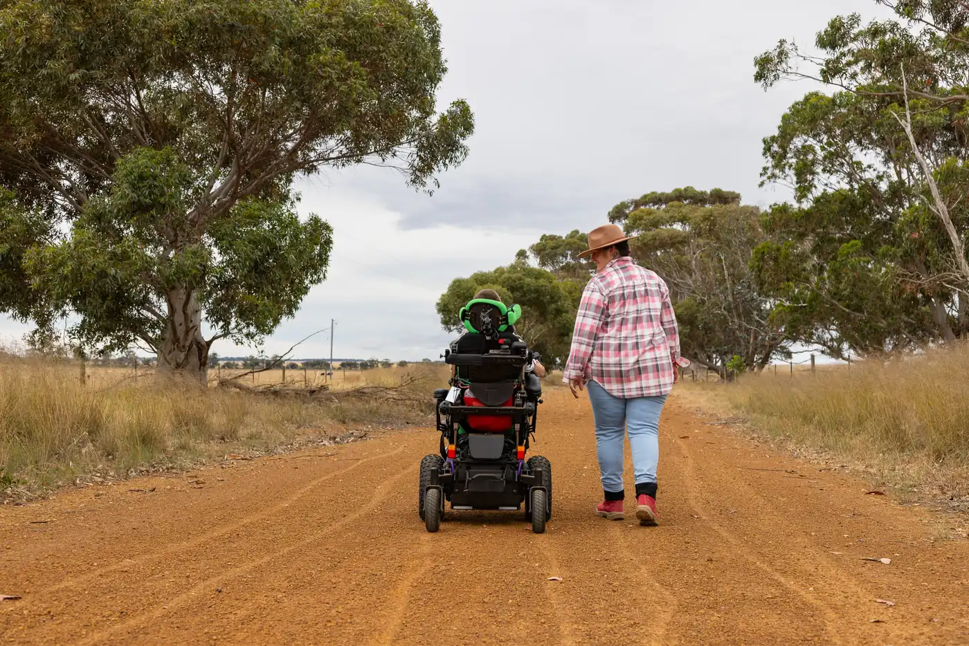 A person in a power wheelchair and a support person walking along a red dirt road